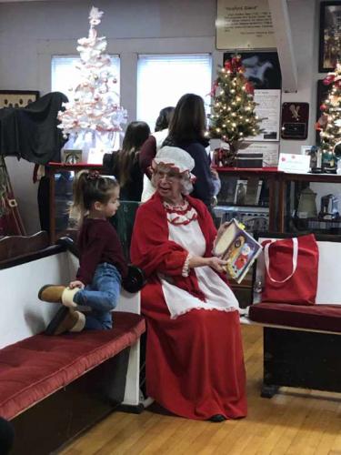 Young girl sitting next to Mrs. Claus who is reading a story to the child.